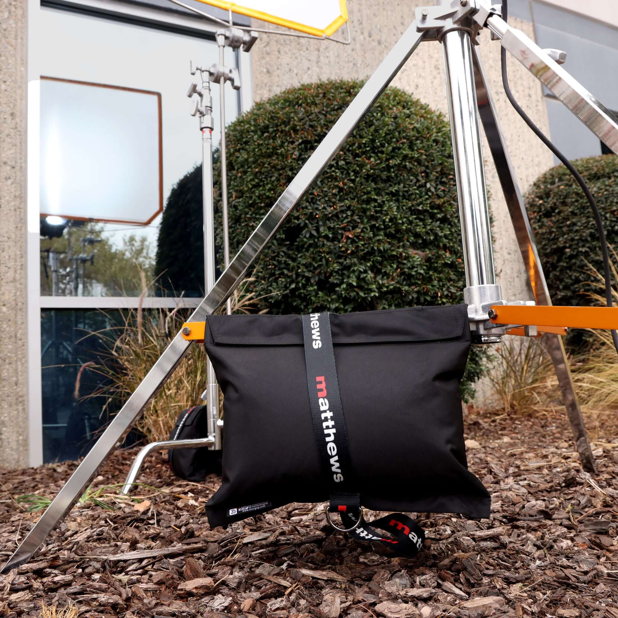 Black sandbag by Matthews securing a lighting stand outdoors, with greenery and building in the background.