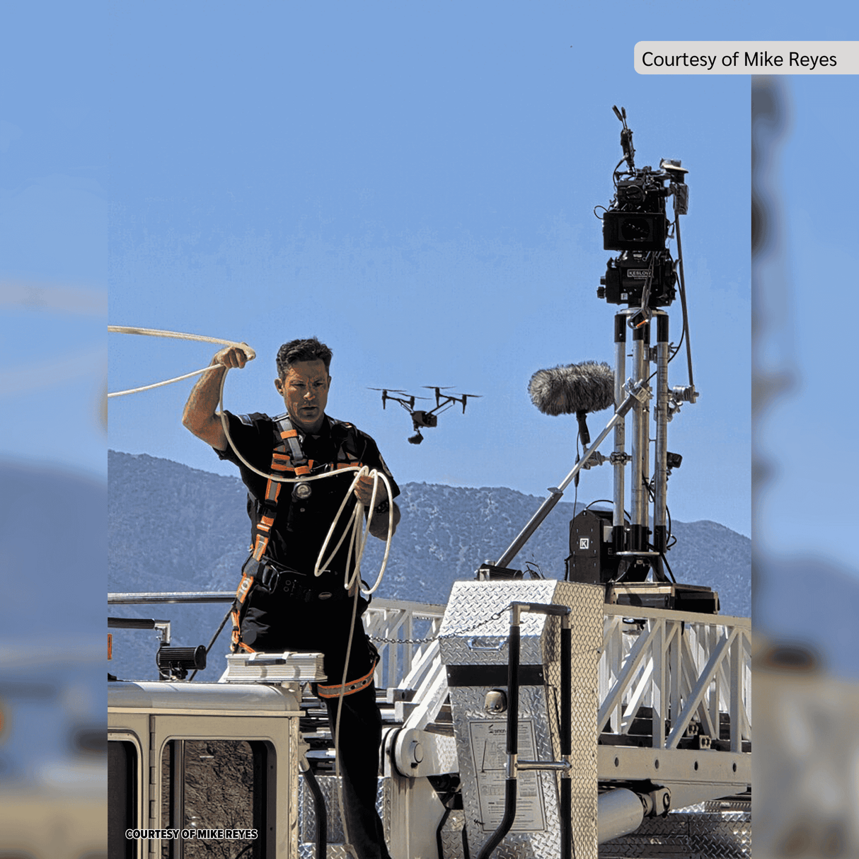 Crew member adjusting equipment on a film set with drone and camera against a mountainous backdrop.