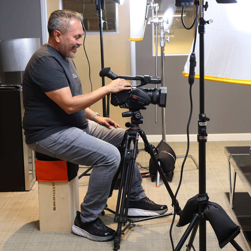 Man sitting on Apple Box Topper™, operating a video camera in a studio setup with lighting equipment.