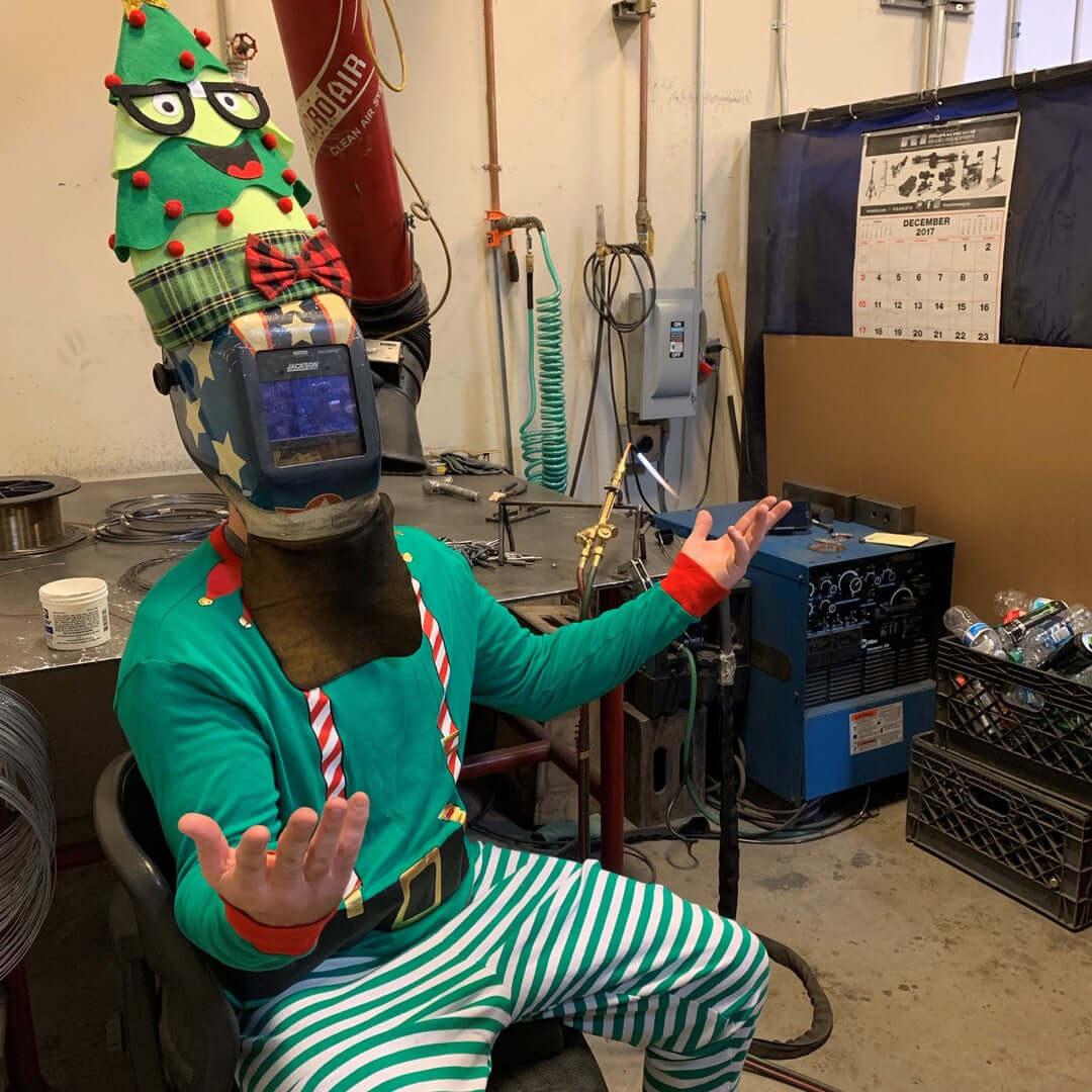 Playful elf in a festive welding helmet and Christmas hat, showcasing holiday spirit in a workshop setting.