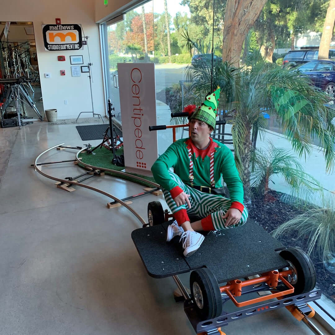 Person dressed as an elf sitting on a cart in a modern studio with a sign for Centipede and greenery in the background.