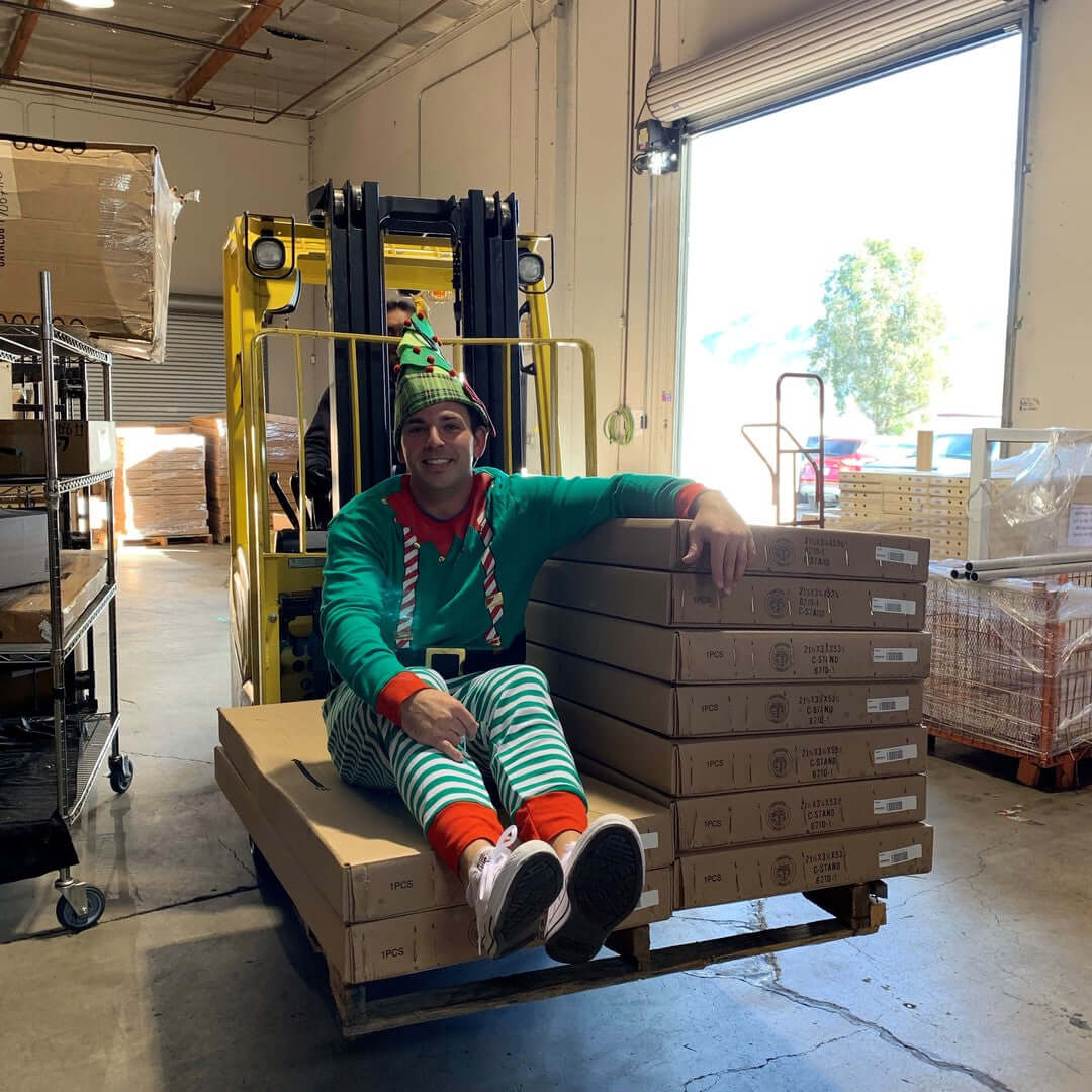 Person in festive elf costume sitting on a pallet of boxes in a warehouse near a forklift.