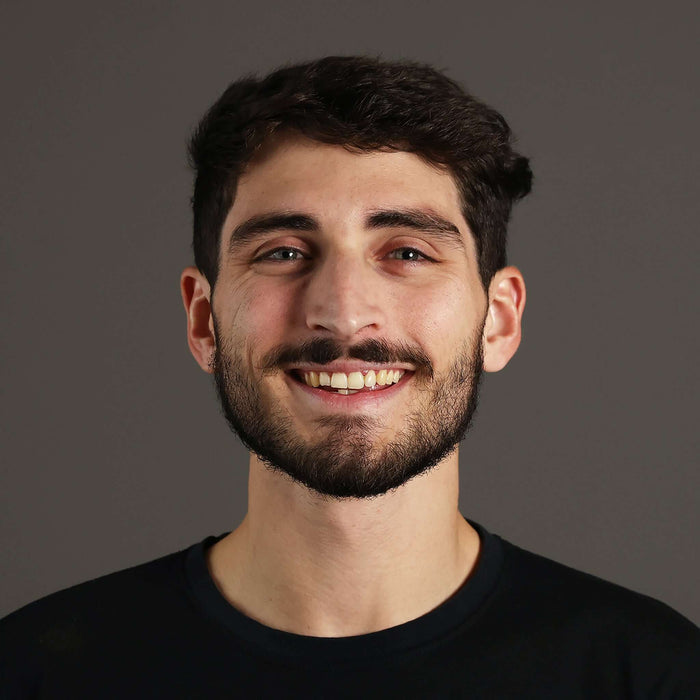 Smiling young man with short dark hair and a beard, wearing a black t-shirt, against a gray background.