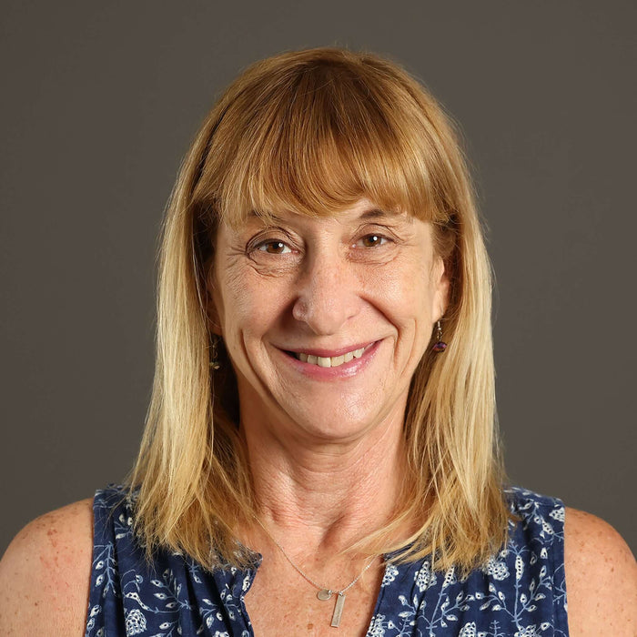 Smiling woman with blonde hair wearing a blue patterned top, set against a gray background.