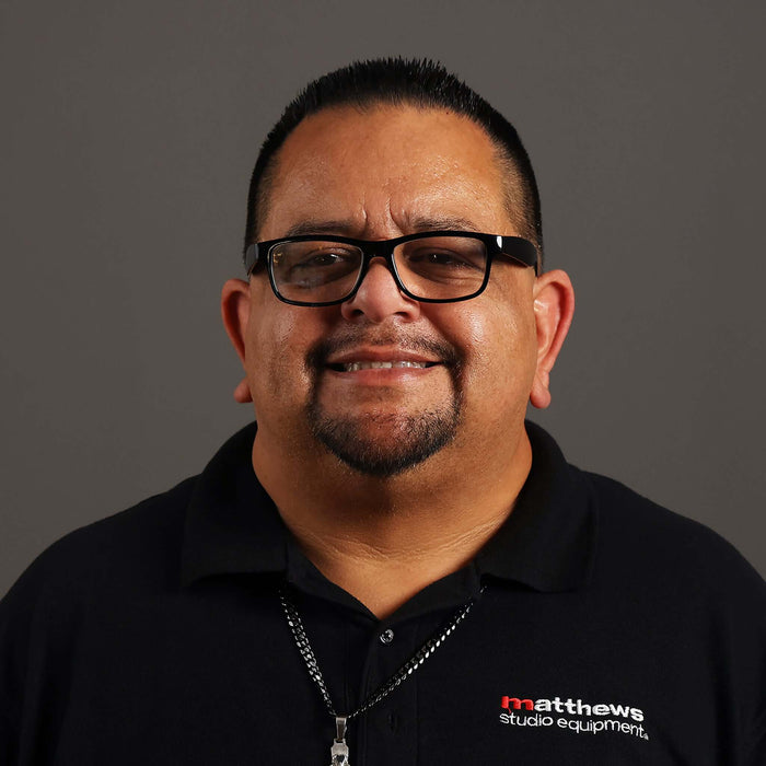 Smiling man in glasses wearing a black shirt with Matthews Studio Equipment logo against a gray background.